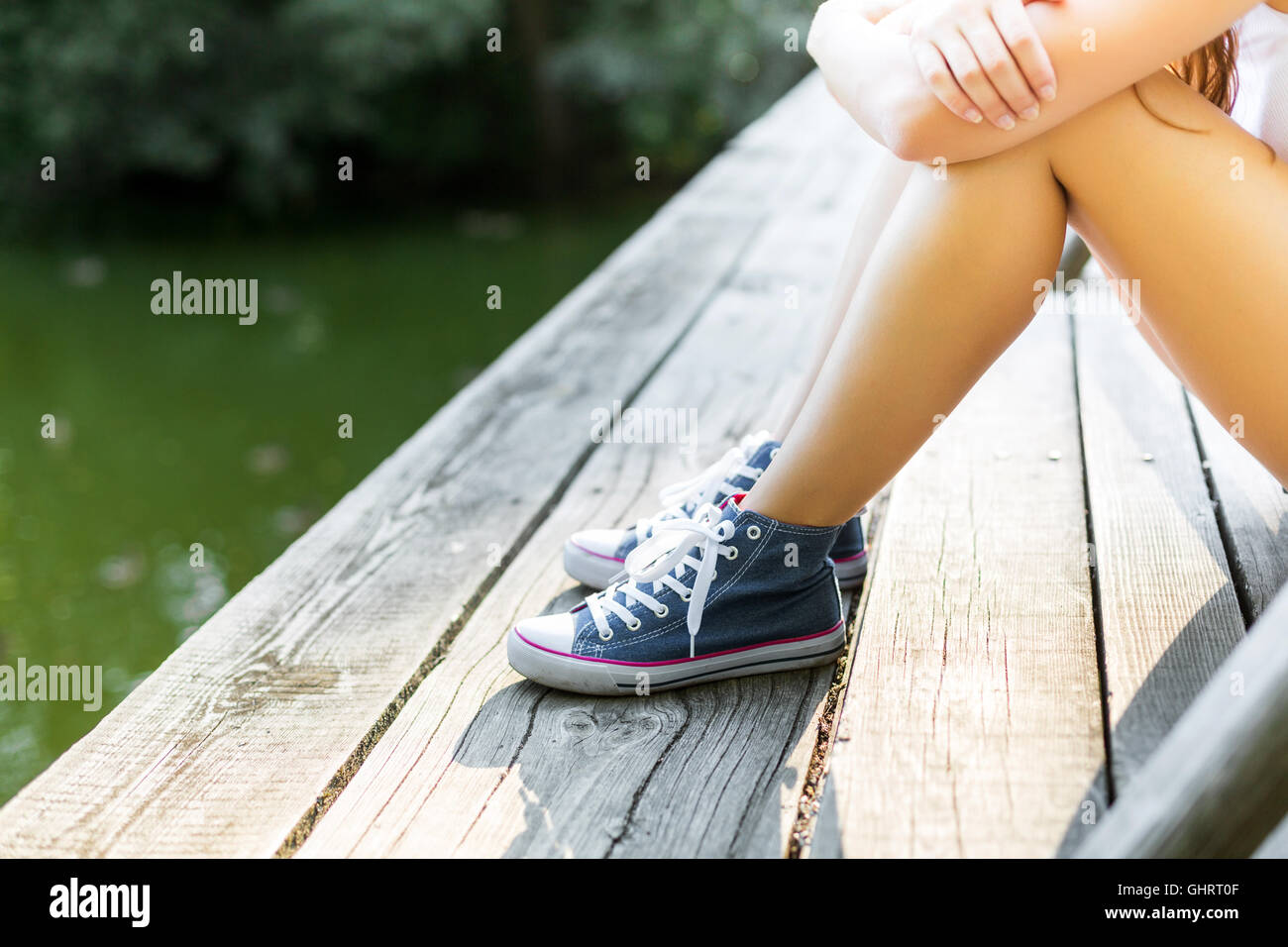 Junge Frau mit schönen sportlichen Beinen sitzen auf einer Holzbrücke Geländer in Jeans Turnschuhen Stockfoto