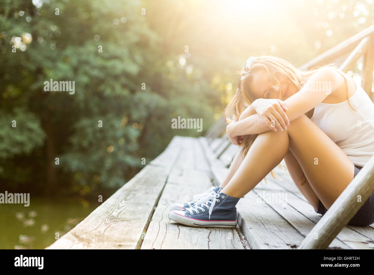 Junge Frau mit schönen sportlichen Beinen sitzen auf einer Holzbrücke Geländer in Jeans Turnschuhen Stockfoto