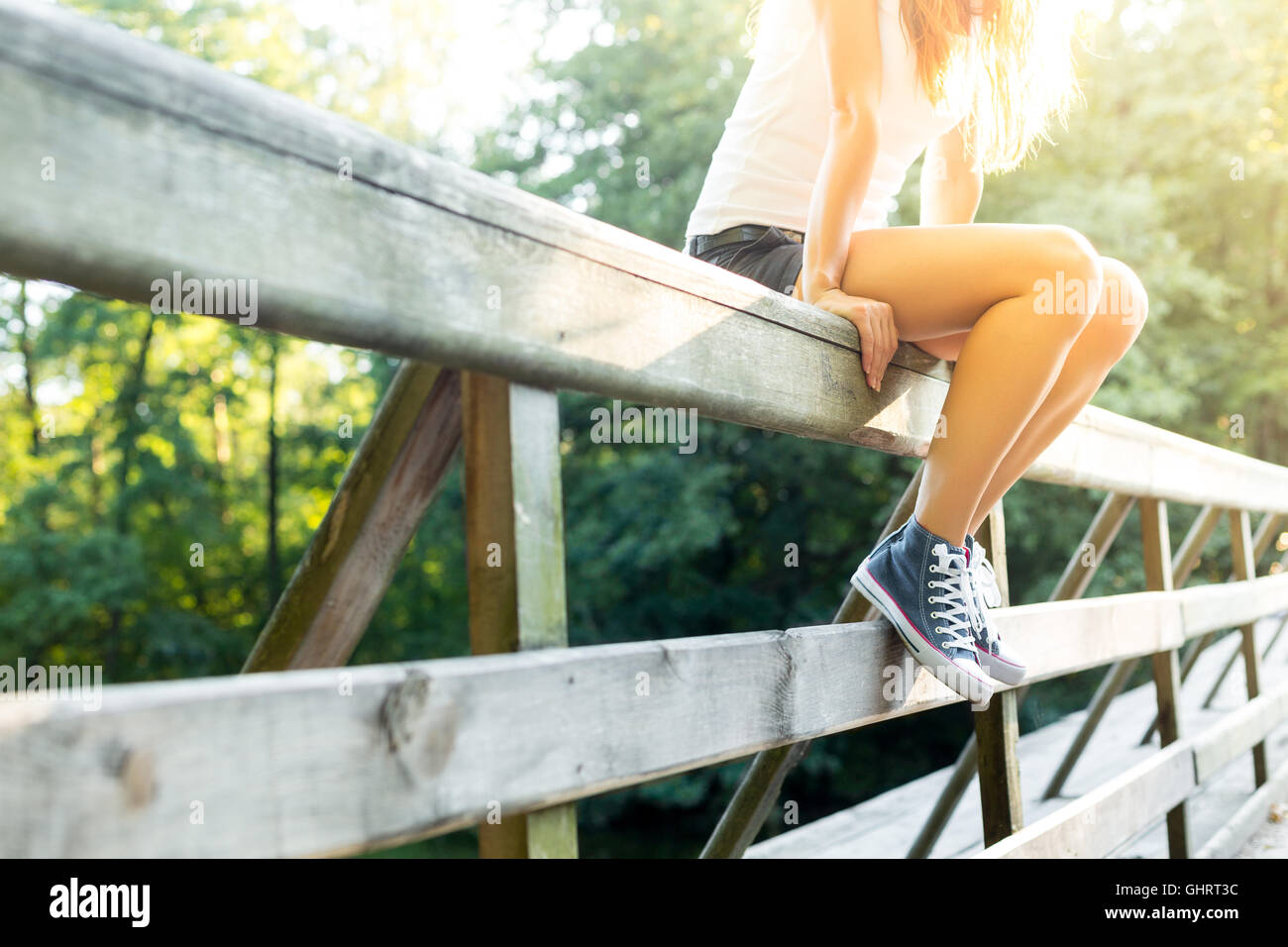 Junge Frau mit schönen sportlichen Beinen sitzen auf einer Holzbrücke Geländer in Jeans Turnschuhen Stockfoto