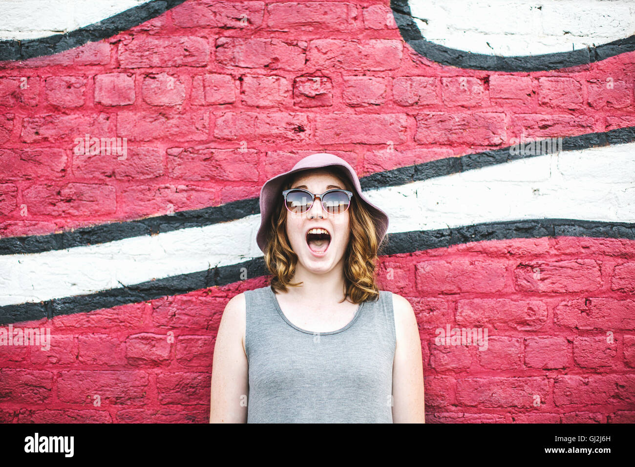 Frau mit Sonnenhut und eine Sonnenbrille vor Wandbild, Mund öffnen Stockfoto