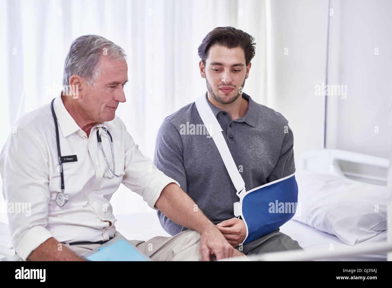 Arzt und Mensch mit Arm in der Schlinge sitzt am Krankenhausbett Stockfoto