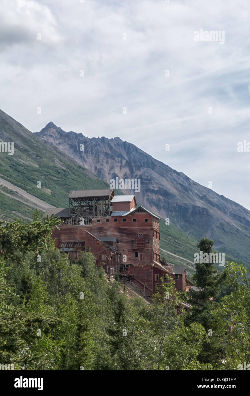 Das verblasste rote Mühle-Haus der Kennicott Mine steht Shorouded von einem Wald mit einem Berg im Hintergrund. Stockfoto