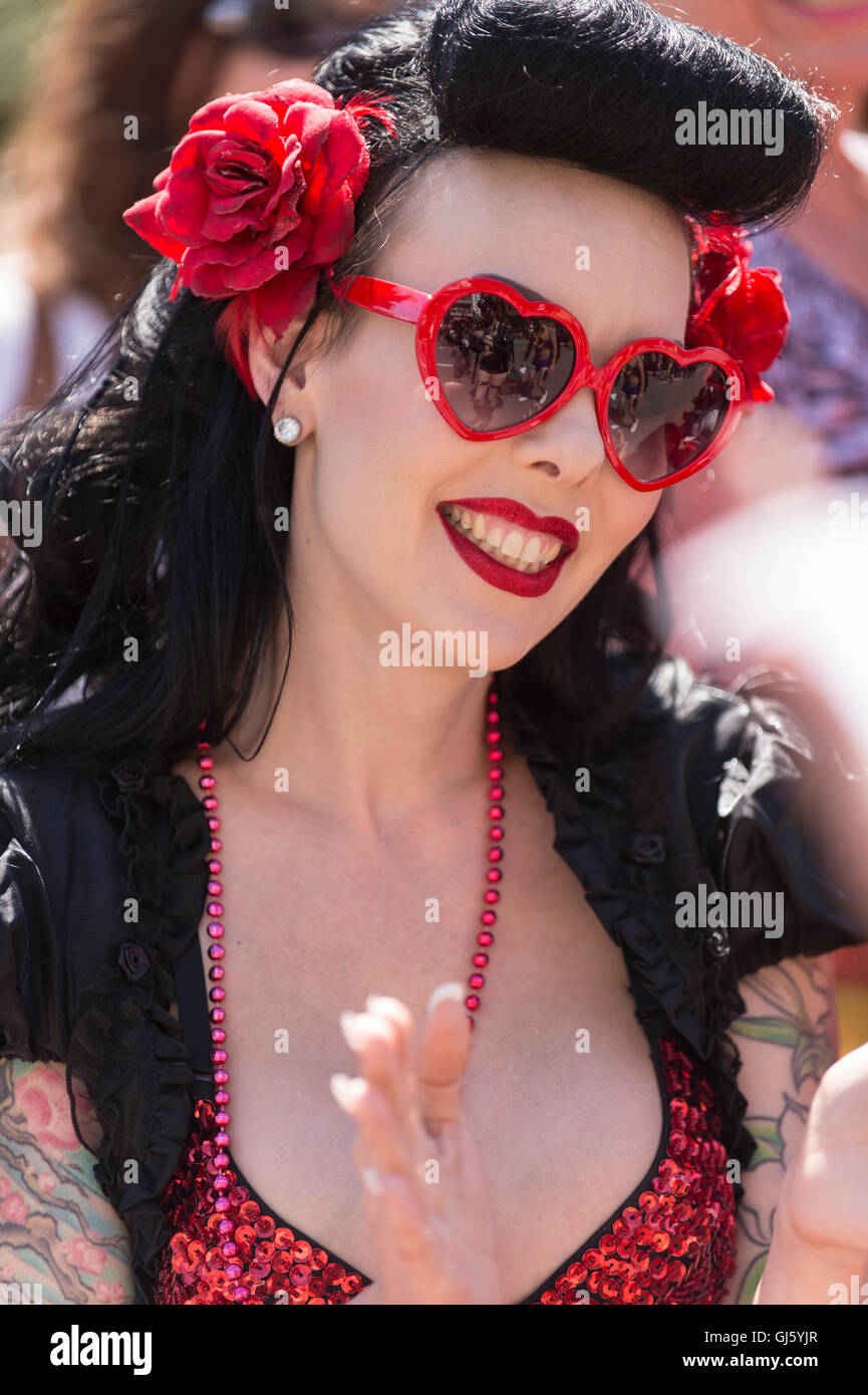 Frau mit schwarzen Haaren, roten Lippen, rote Blumen in ihrem Haar und roten Herzen geformt Sonnenbrille, lächelnd. Stockfoto