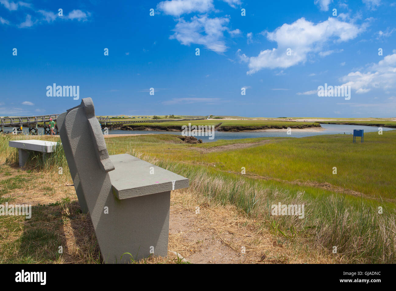 Konkrete Bank in der Nähe der Gehweg zu den Dünen. Holzsteg erstreckt sich über Marschland gegenüber den weit entfernten Dünen und das Meer In Sandwi Stockfoto