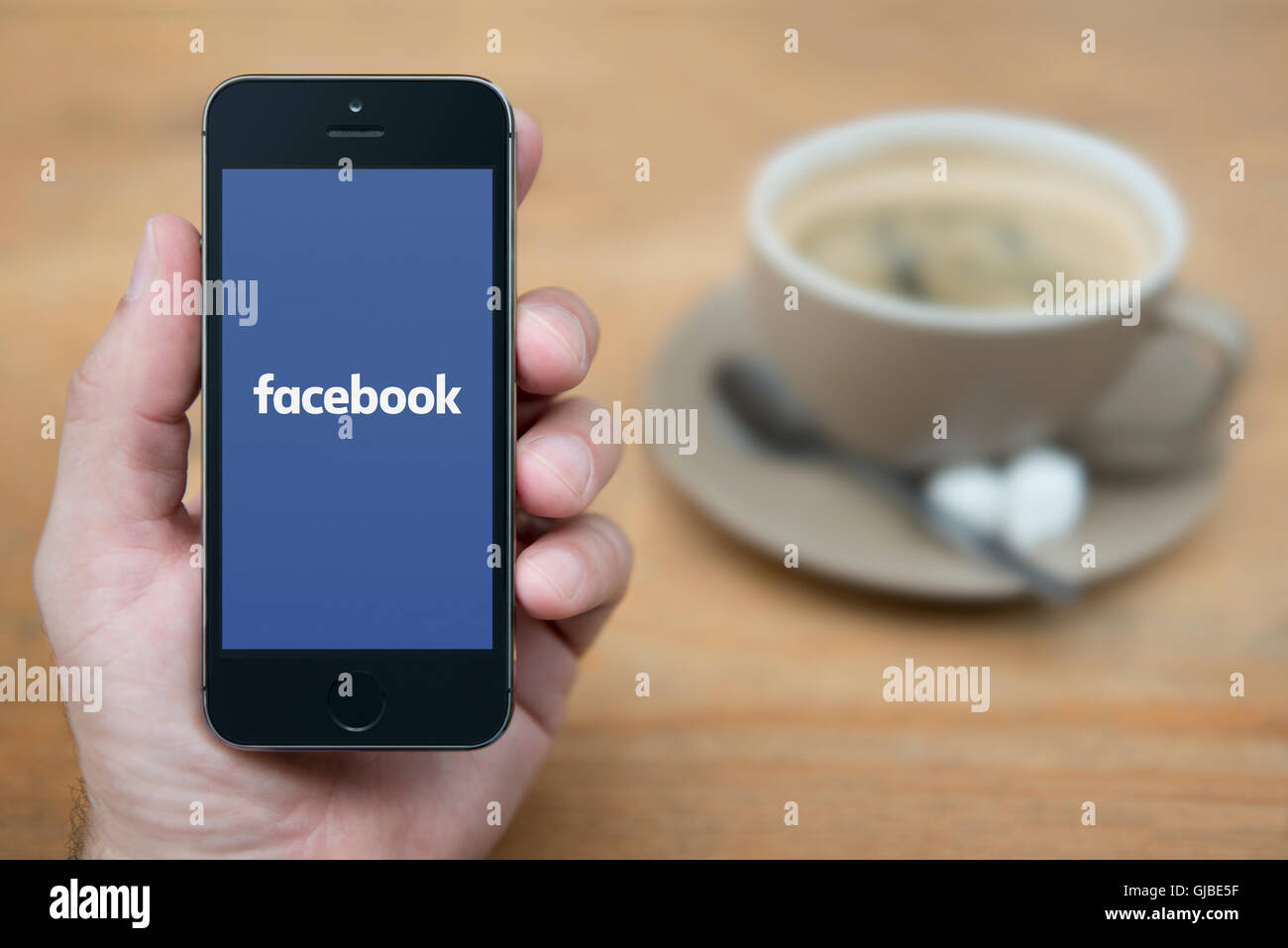 Ein Mann schaut auf seinem iPhone die das Facebook-Logo anzeigt, während bei einer Tasse Kaffee (nur zur redaktionellen Verwendung) saß. Stockfoto