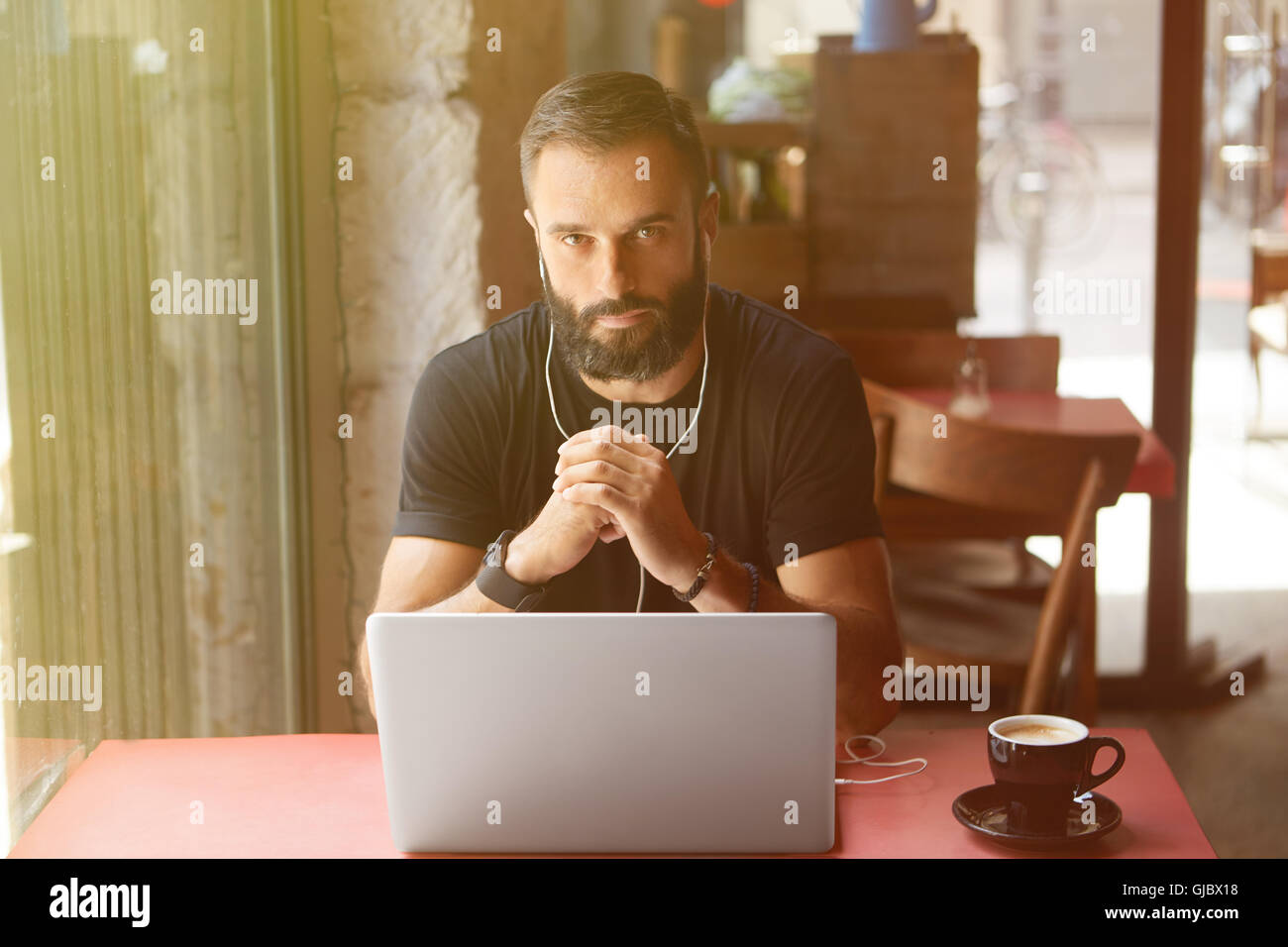 Junge Bearded Geschäftsmann tragen schwarz Tshirt arbeiten Laptop Urban Cafe.Man sitzen Holz Tisch Tasse Kaffee hören Music.Coworking Prozess Business Startup.Blurred Background.Sunlight Wirkung. Stockfoto
