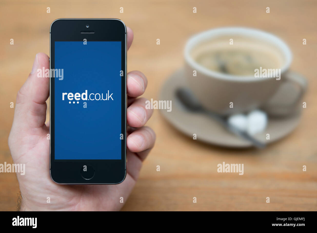Ein Mann schaut auf seinem iPhone die das Reed-Rekrutierung-Logo anzeigt, während mit einer Tasse Kaffee (nur zur redaktionellen Verwendung) saß. Stockfoto