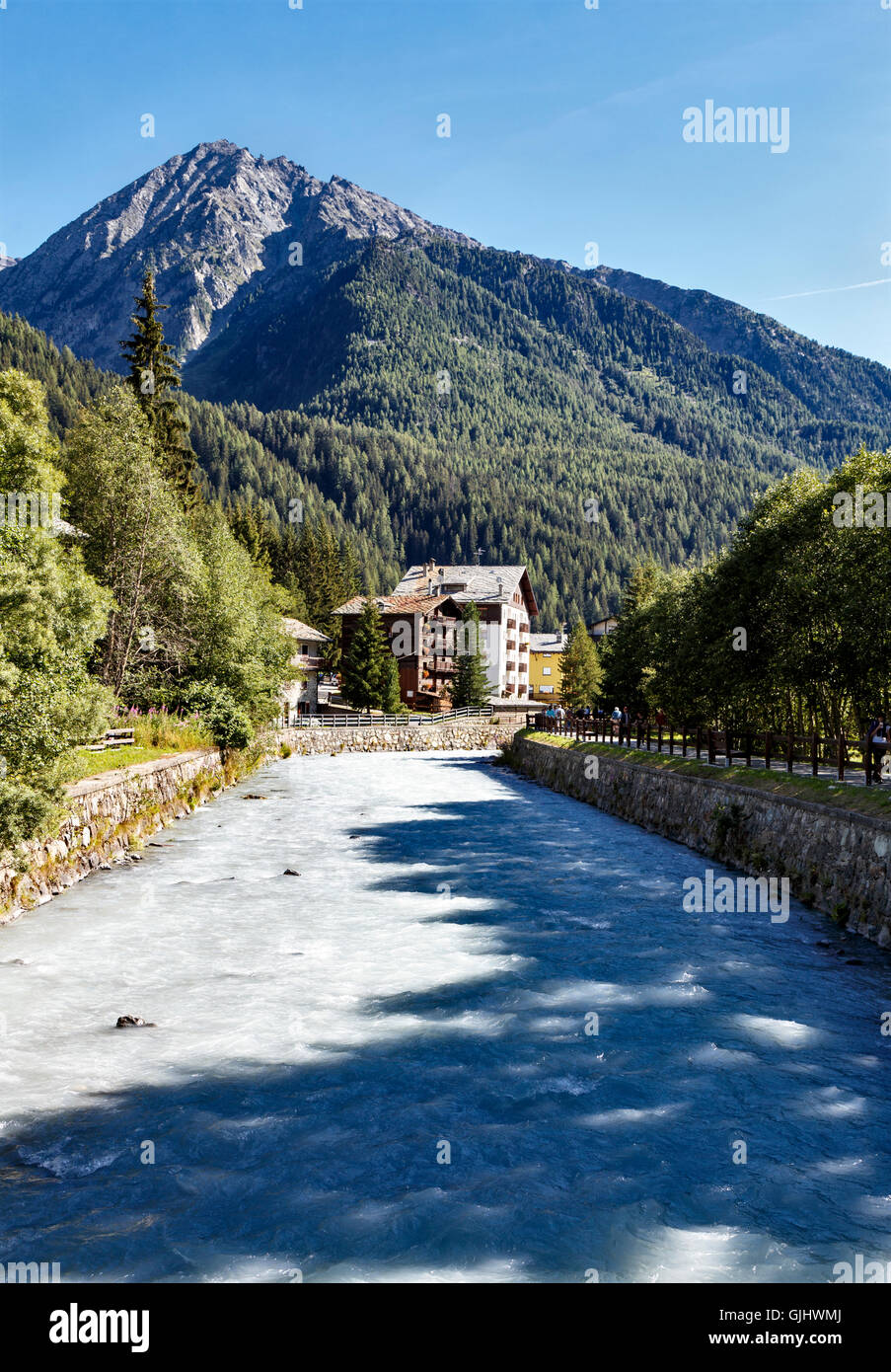 Fluss, der durch eine steile bewaldete Tal mit traditionellen Häusern unter einem hohen Berggipfel in Champoluc, nördlichen ich Stockfoto
