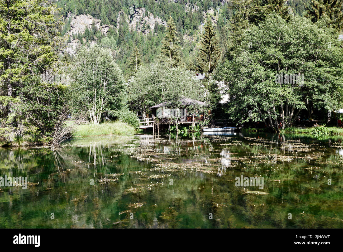 Ruhigen Bergsee in Champoluc, Italien ein Holzchalet mit kleinen Boot unter steilen bewaldeten Berghängen Stockfoto