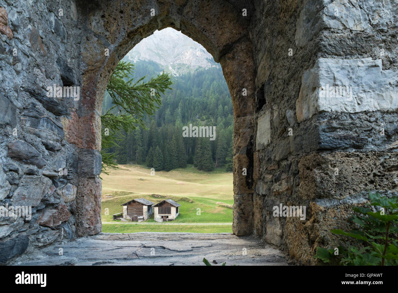 Blick durch das leere Tor Bogen einer alten Burgruine auf zwei Hütten. Grünen Wald und Wiese im Hintergrund sichtbar. Stockfoto