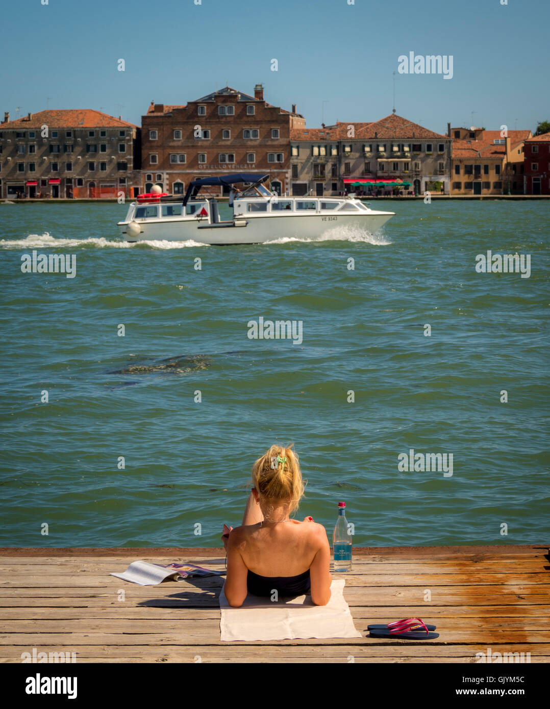 Junge Frau liegend auf einem Holzsteg über den Canale della Giudecca, mit einem Boot und in der Ferne die Insel Giudecca. V Stockfoto