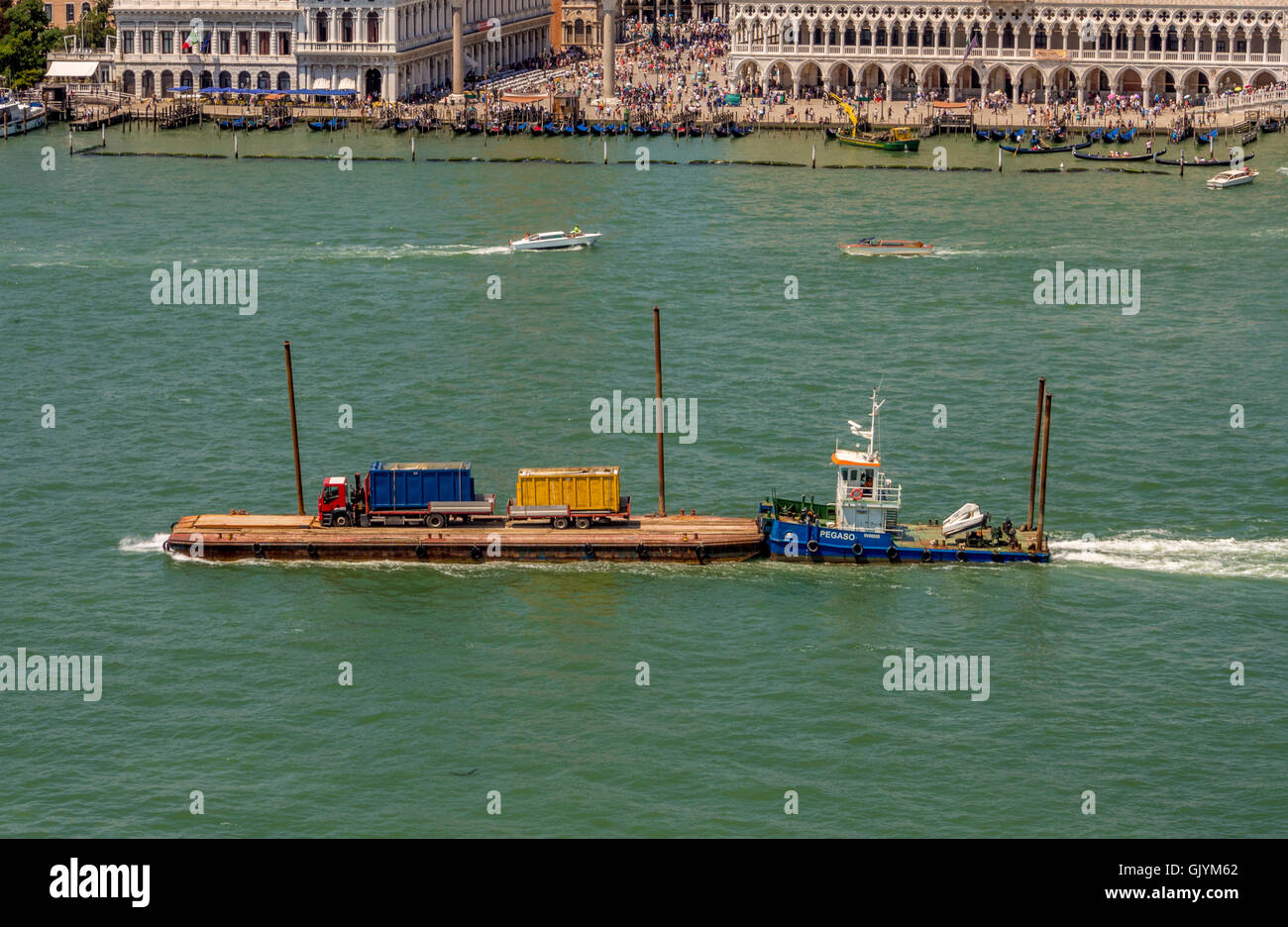LKW und Anhänger auf einem Lastkahn von einem Schlepper am Markusplatz Becken mit Dogenpalast in der Ferne geschoben. Venedig, Italien. Stockfoto