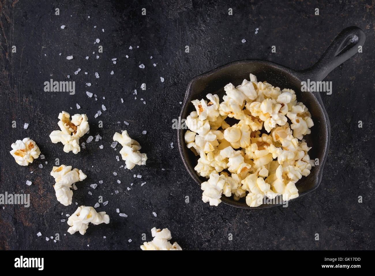 Vorbereitete gesalzene Popcorn serviert mit Meersalz in kleinen Eisen-Guss-Pfanne über schwarzes Eisen strukturiertem Hintergrund. Draufsicht mit Kopie spac Stockfoto