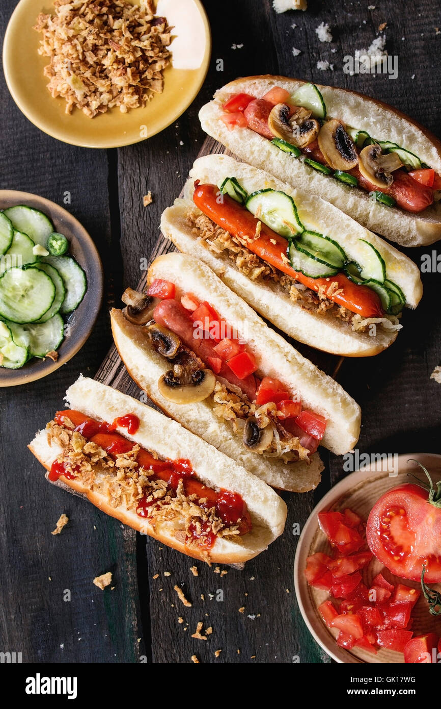 Auswahl an hausgemachten Hot Dogs mit Wurst, gebratenen Zwiebeln, Tomaten und Gurken, serviert mit Zutaten in verschiedenen Platten auf Stockfoto