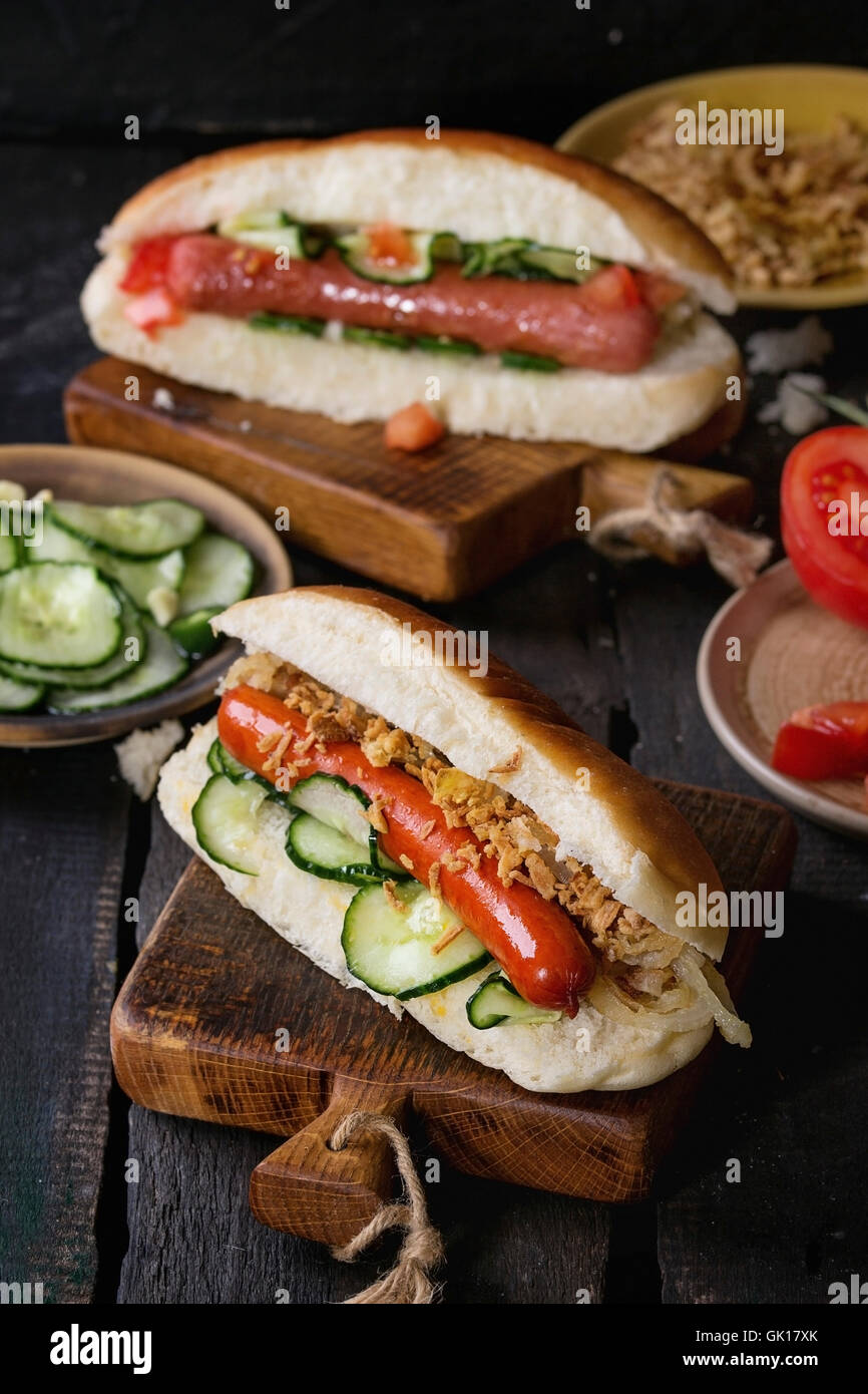 Zwei hausgemachte Hot Dogs mit Wurst, gebratenen Zwiebeln, Tomaten und Gurken, serviert mit Zutaten in verschiedenen Platten auf Holz c Stockfoto
