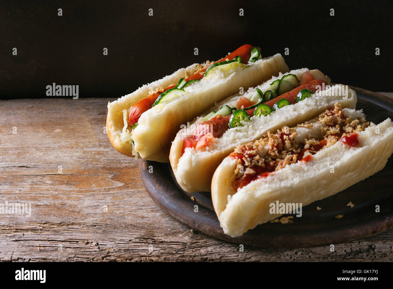 Auswahl an hausgemachten Hot Dogs mit Wurst, gebratenen Zwiebeln, Tomaten und Gurken, serviert auf Holz Schneidebrett über alte hölzerne Stockfoto