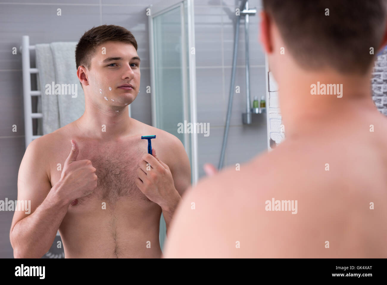 Zufrieden, junger Mann mit Schnitten auf seinem Gesicht ein Rasiermesser stehend vor dem Spiegel in das modern geflieste Bad zu Hause hält. Stockfoto