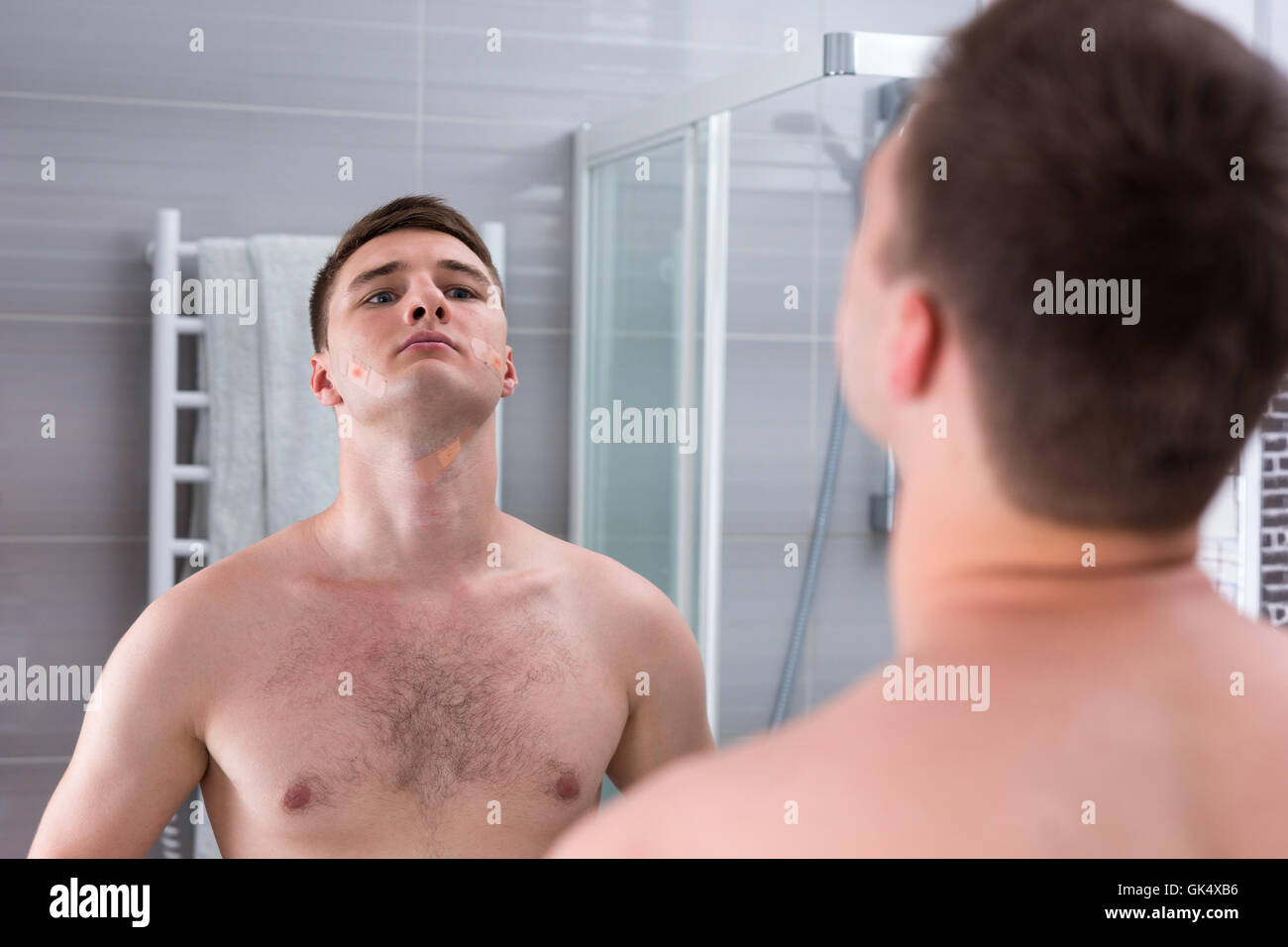 Junger Mann nach der schlechten Rasur mit Pflaster auf seinem Gesicht stand vor dem Spiegel in das modern geflieste Bad zu Hause Stockfoto