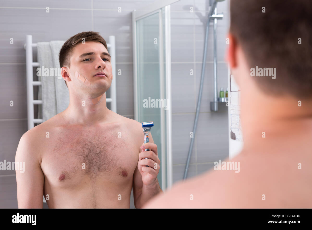 Gut aussehender Mann nach der schlechten Rasur mit Pflaster auf seinem Gesicht ein Rasiermesser und stand vor dem Spiegel in das modern geflieste Bad zu Hause hält. Stockfoto