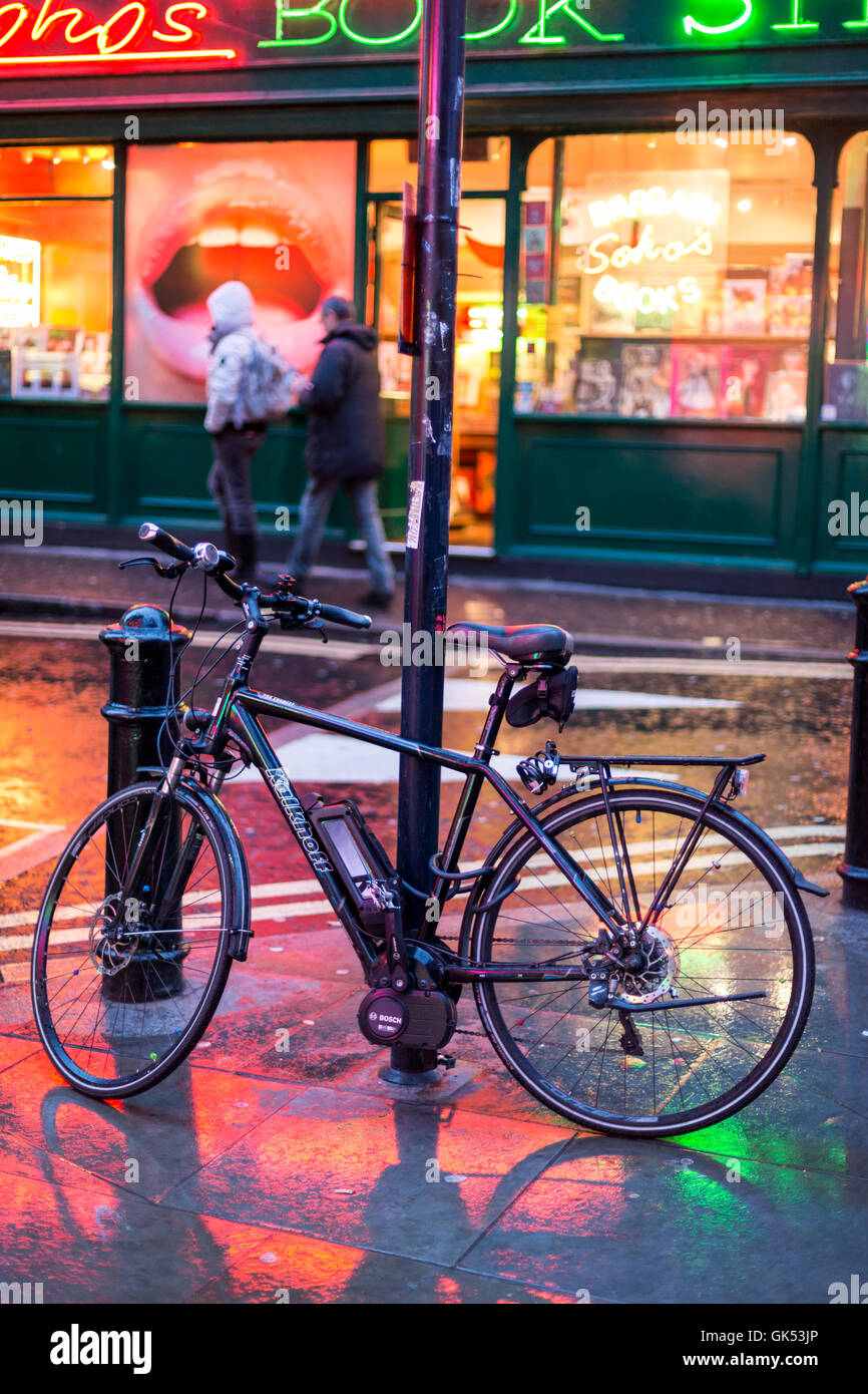 Eine elektrische Lenkhilfe Fahrrad ist zu einem Wegweiser außerhalb ein Buch Shop in Londons Stadtteil Soho gesperrt. Nässe widerspiegeln, bunten Leuchtreklamen Stockfoto