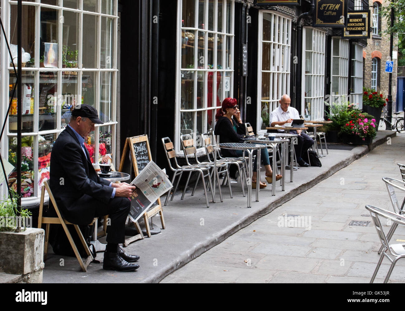 Ein älterer Mann liest eine Zeitung zusammen mit anderen Einheimischen und genießt Getränke und Erfrischungen an den Tischen im Freien in kleinen Cafés Stockfoto