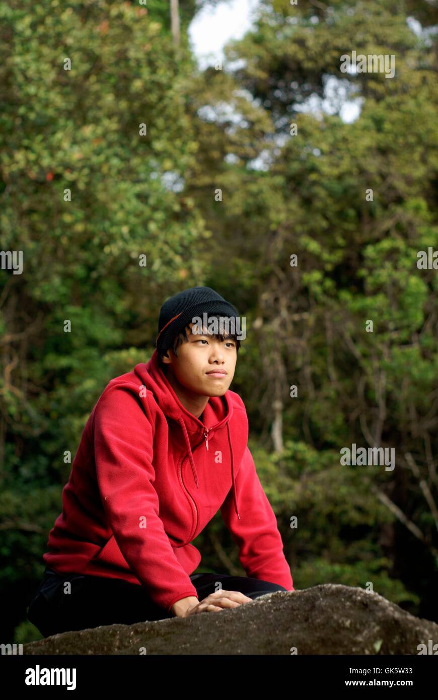 Asiatischen Mann kniet auf Felsen im Freien tragen kalte Kleidung weit wegschauen Stockfoto