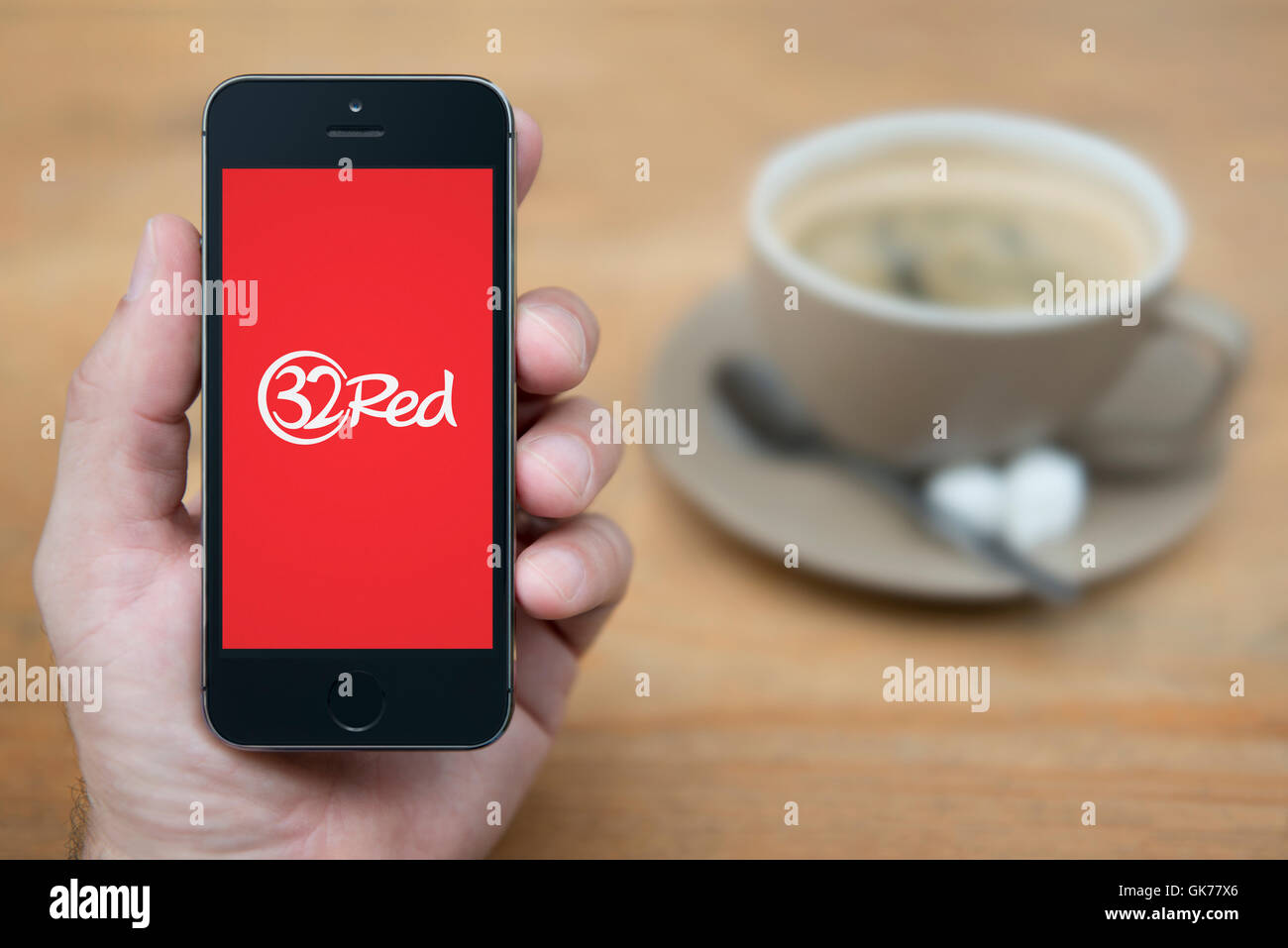Ein Mann schaut auf seinem iPhone die das 32 rote Logo anzeigt, während mit einer Tasse Kaffee (nur zur redaktionellen Verwendung) saß. Stockfoto