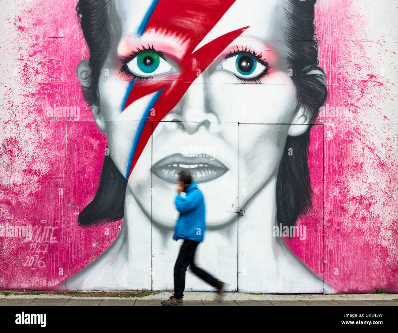 Newcastle Upon Tyne, England, Vereinigtes Königreich. 19. August 2016. Wetter: Ein Farbtupfer an einem grauen Freitagmorgen in Gateshead als Mann telefonieren mit Handy geht vorbei an ein riesiges Wandgemälde von David Bowie in der Nähe von The Sage Gateshead. Eine Bande von Regen wird voraussichtlich um am Freitag in ganz Großbritannien zu fegen. Bildnachweis: Alan Dawson News/Alamy Live-Nachrichten Stockfoto