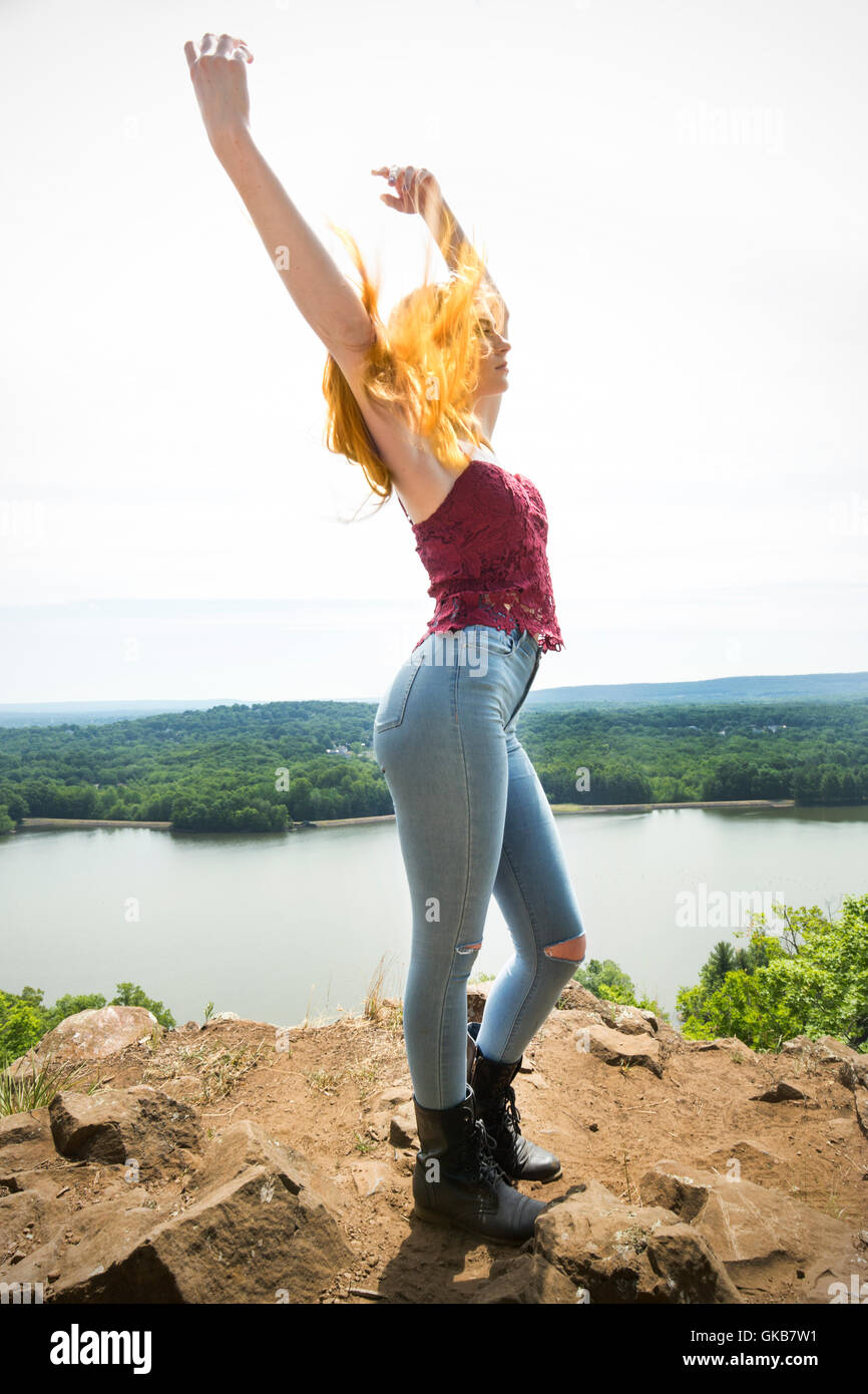 Schöne rote Kopf in Röhrenjeans, schwarze Stiefel und rote oben am Berg oben mit Wind im Haar, erhobenen Armen und einem See unten. Stockfoto