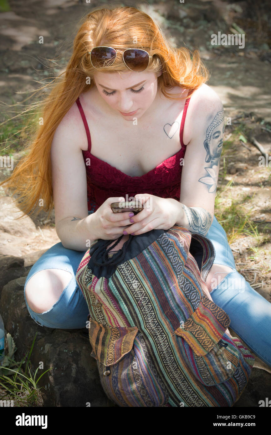 Schöne rote Kopf Frau in Röhrenjeans und rotes Top, sitzt auf dem Boden im Wald, Blick auf ein Handy. Stockfoto
