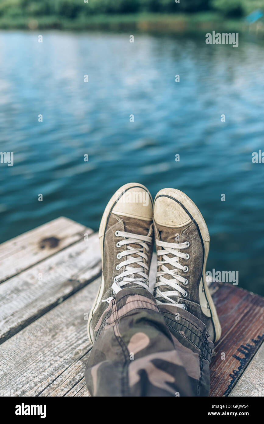 Mann mit gekreuzten Beinen am Flussufer Pier, lässige junge Mann tragen Turnschuhe, die durch den Fluss im Sommernachmittag sitzen entspannen Stockfoto