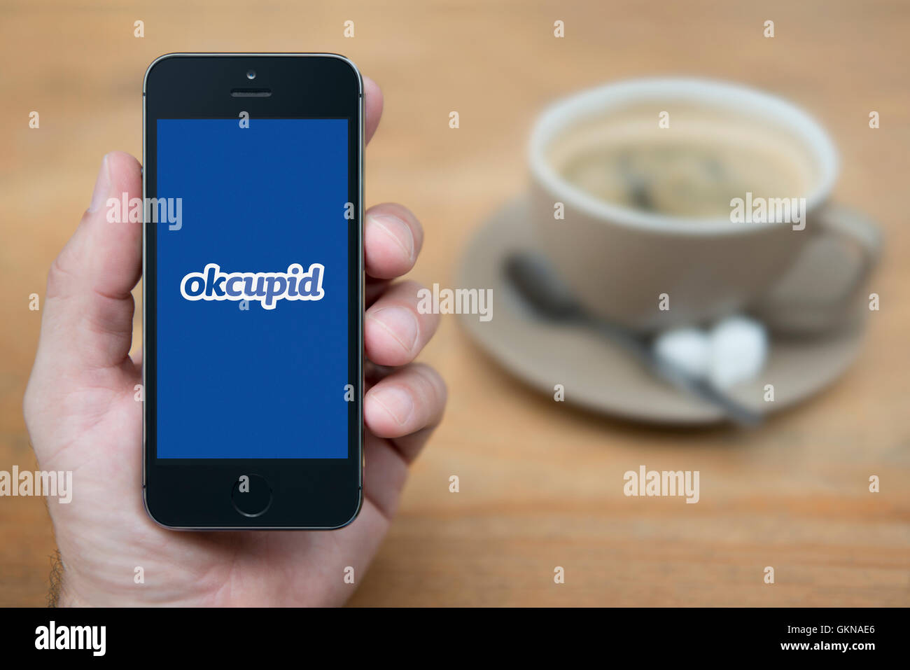 Ein Mann schaut auf seinem iPhone das OK Cupid-Logo anzeigt, während bei einer Tasse Kaffee (nur zur redaktionellen Verwendung) saß. Stockfoto