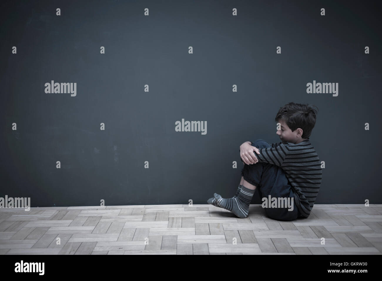 Junge sitzt am Boden gegen die graue Wand umarmt Knie Stockfoto