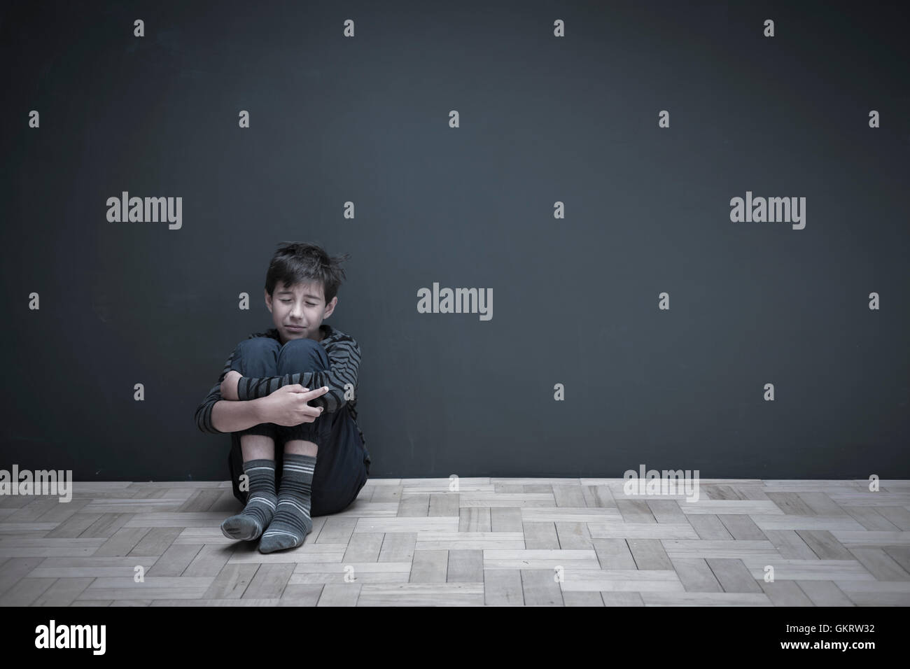 Junge sitzt am Boden gegen die graue Wand umarmt Knie Stockfoto