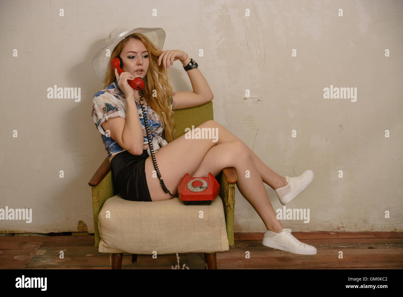 hübsche Blondine in weißen Hut sitzt auf dem Sessel mit gekreuzten Beinen kommunizieren per Telefon und leicht räumt Haar Stockfoto