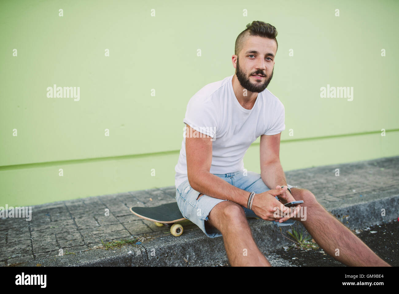 Porträt des Mannes mit Smartphone sitzen auf seinem Skateboard an carb Stockfoto