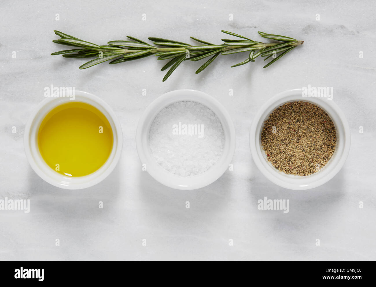 Draufsicht der Schüssel mit Olivenöl, Salz, Pfeffer und Rosmarin auf Marmortisch Stockfoto
