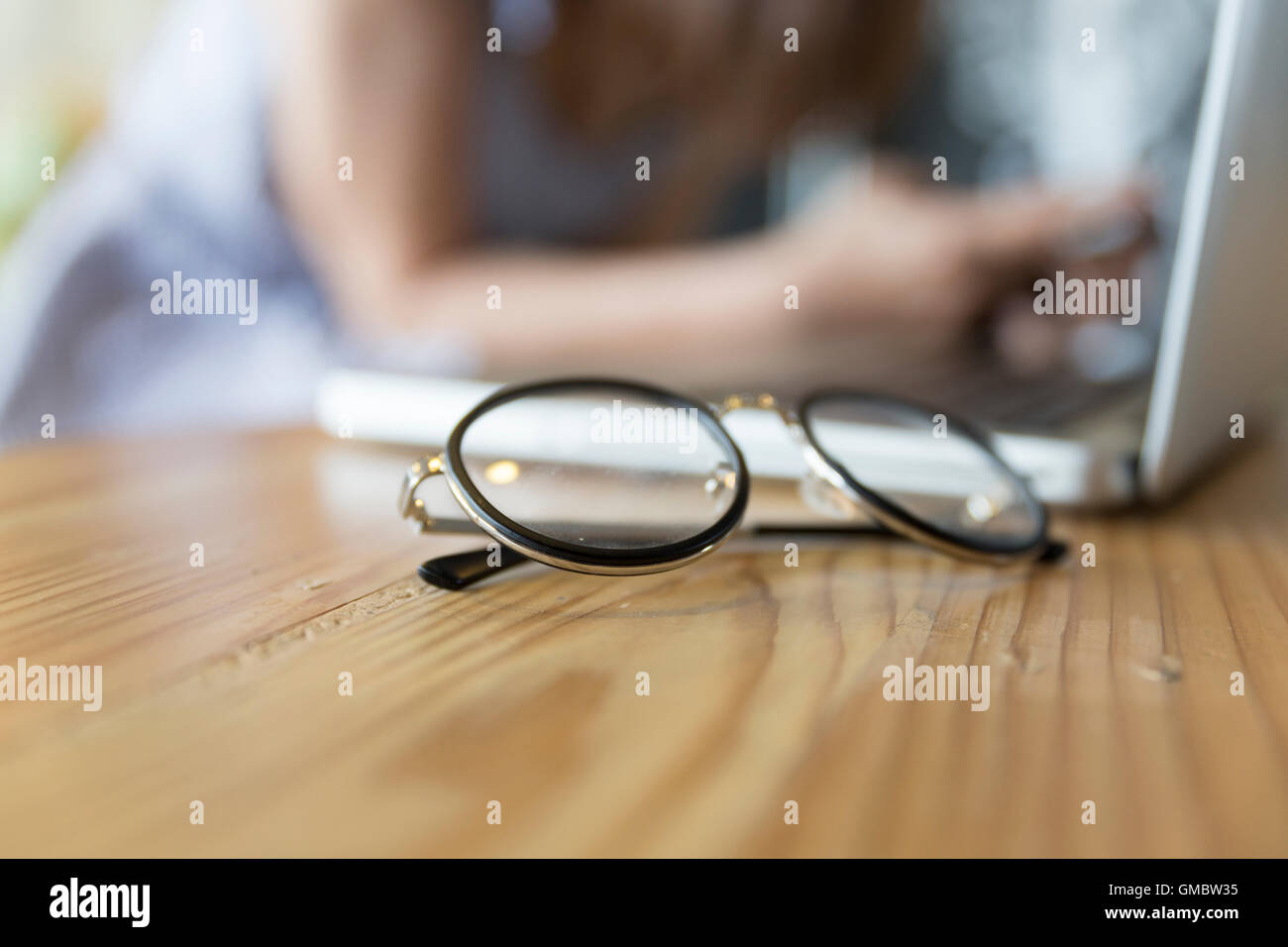 Brillen mit Laptop Computer Notebook am Schreibtisch aus Holz mit Hintergrund der Frau auf Stuhl sitzend Stockfoto