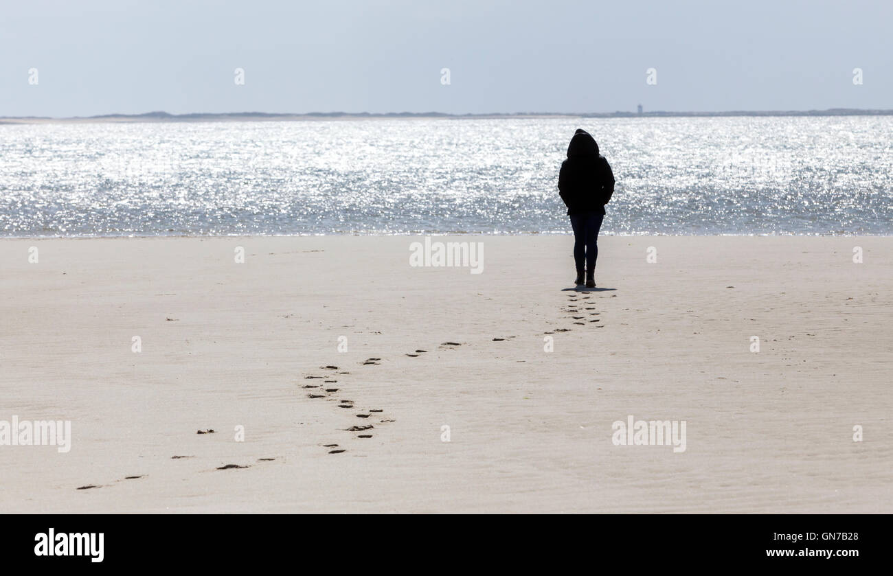 Eine Silhouette einer jungen Frau steht am Strand, so dass ihre Fussspuren im Sand und ein Leuchtturm in der Ferne. Stockfoto