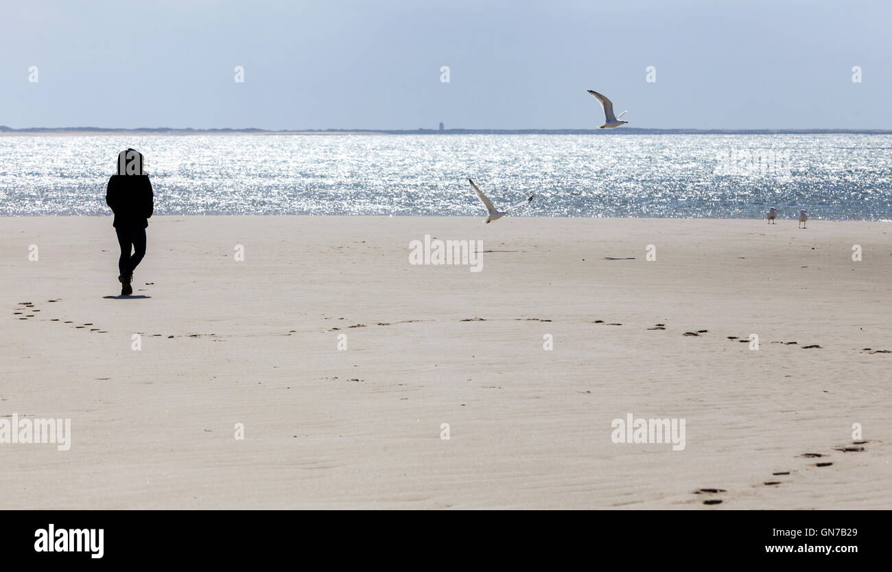 Eine Silhouette einer jungen Frau steht am Strand, so dass ihre Fussspuren im Sand und ein Leuchtturm in der Ferne. Stockfoto