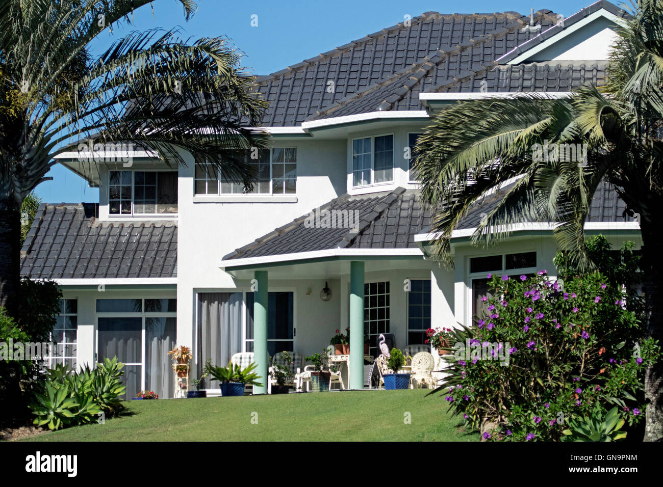 Moderne luxuriöse zweistöckiges Haus mit schwarz gefliest Dach, weiße Wände, Veranda, Garten mit Rasen, blühenden Sträuchern, Palmen, Stockfoto