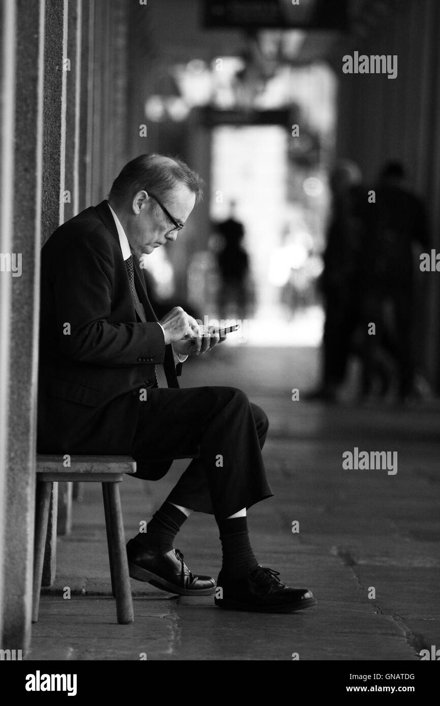 Ein reifer Mann sitzt auf einem Sitz außerhalb Geschäfte Blick auf sein Handy. Stockfoto