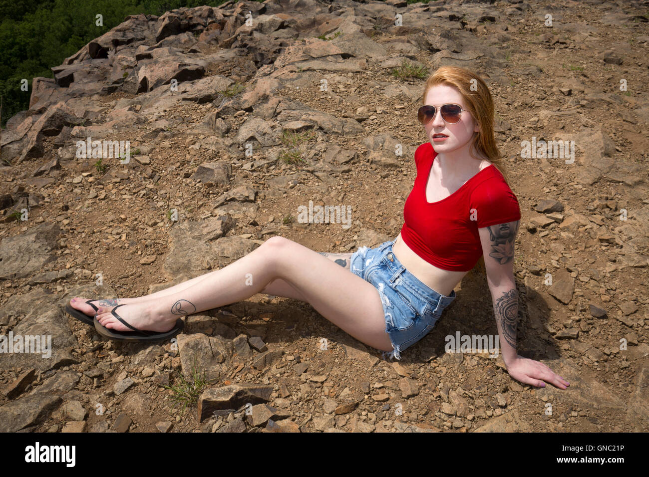 Schöne Frau mit langen roten Haaren, in Jeans-Shorts und rote Hemd, sitzen auf den Felsen des Berggipfel im Sommer. Stockfoto