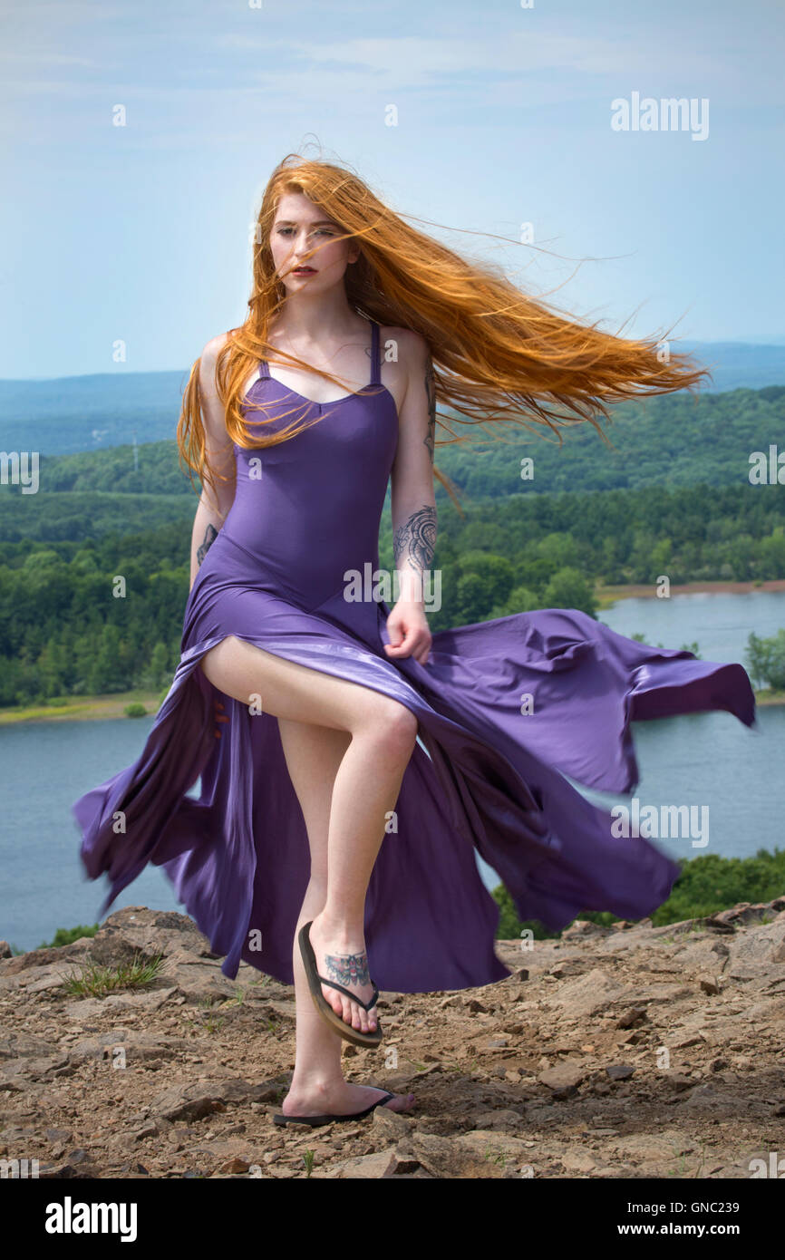 Dramatische Porträt der schönen roten Kopf Frau in fließenden lila Kleid, auf Berggipfel mit Wind im Haar und einem See unten. Stockfoto