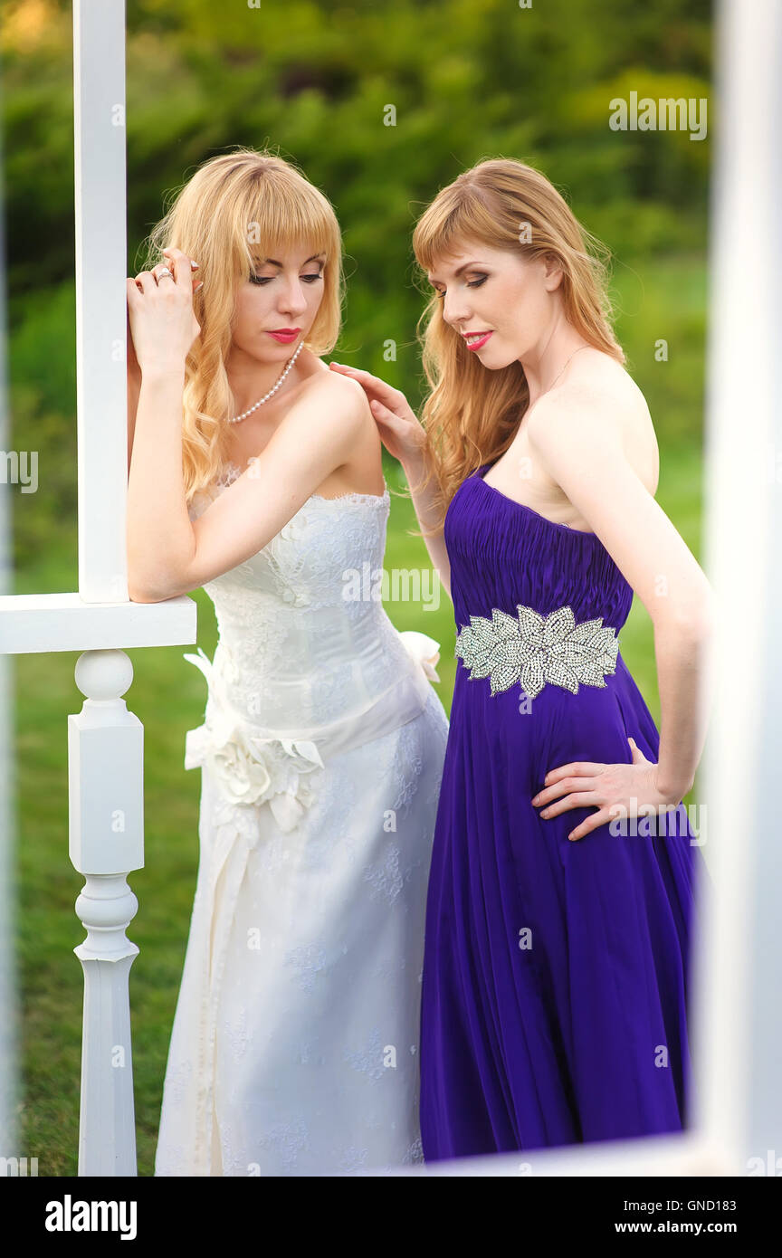 Braut und Brautjungfer außerhalb. Zwei Mädchen posiert auf der grünen Wiese. Braut im Hochzeitskleid. Brautjungfer in einem lila Abendkleid. Stockfoto