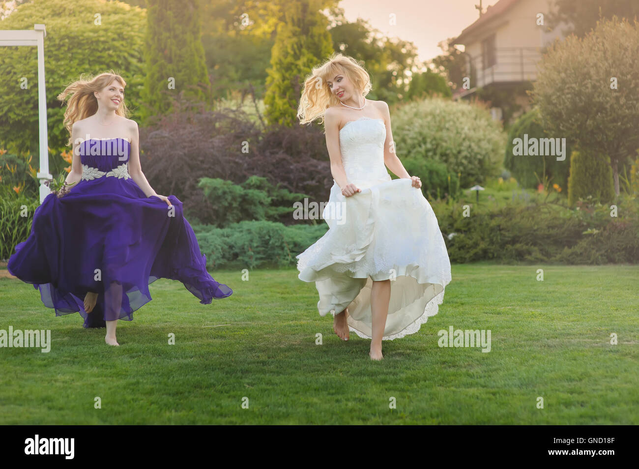 Braut und Brautjungfer außerhalb. Zwei schöne Mädchen auf Rasen laufen. Braut im Hochzeitskleid. Brautjungfer in einem lila Abendkleid. Stockfoto