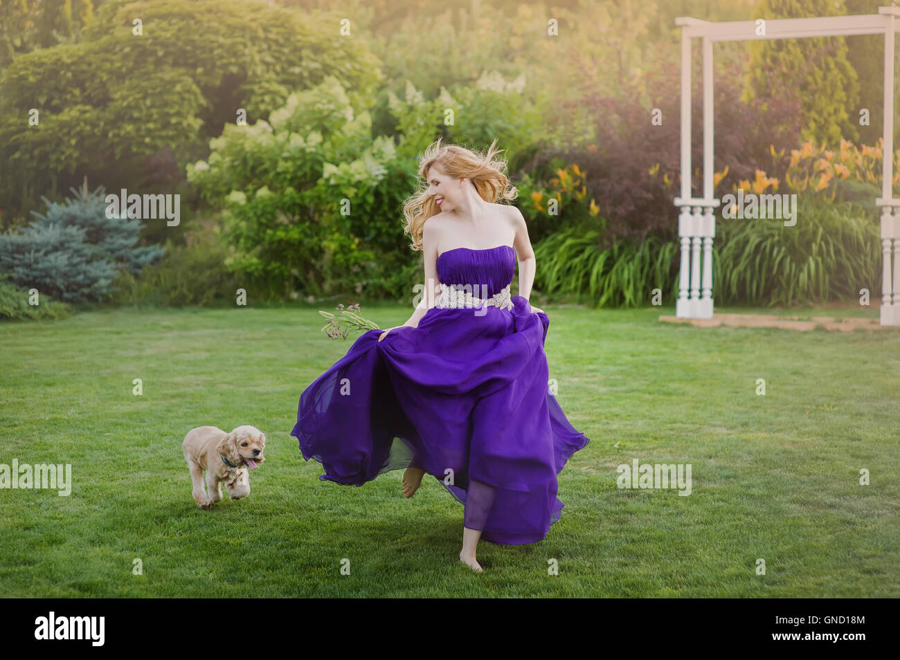 Schöne Mädchen auf Rasen mit Hund laufen. Brautjungfer in einem lila Abendkleid. Stockfoto