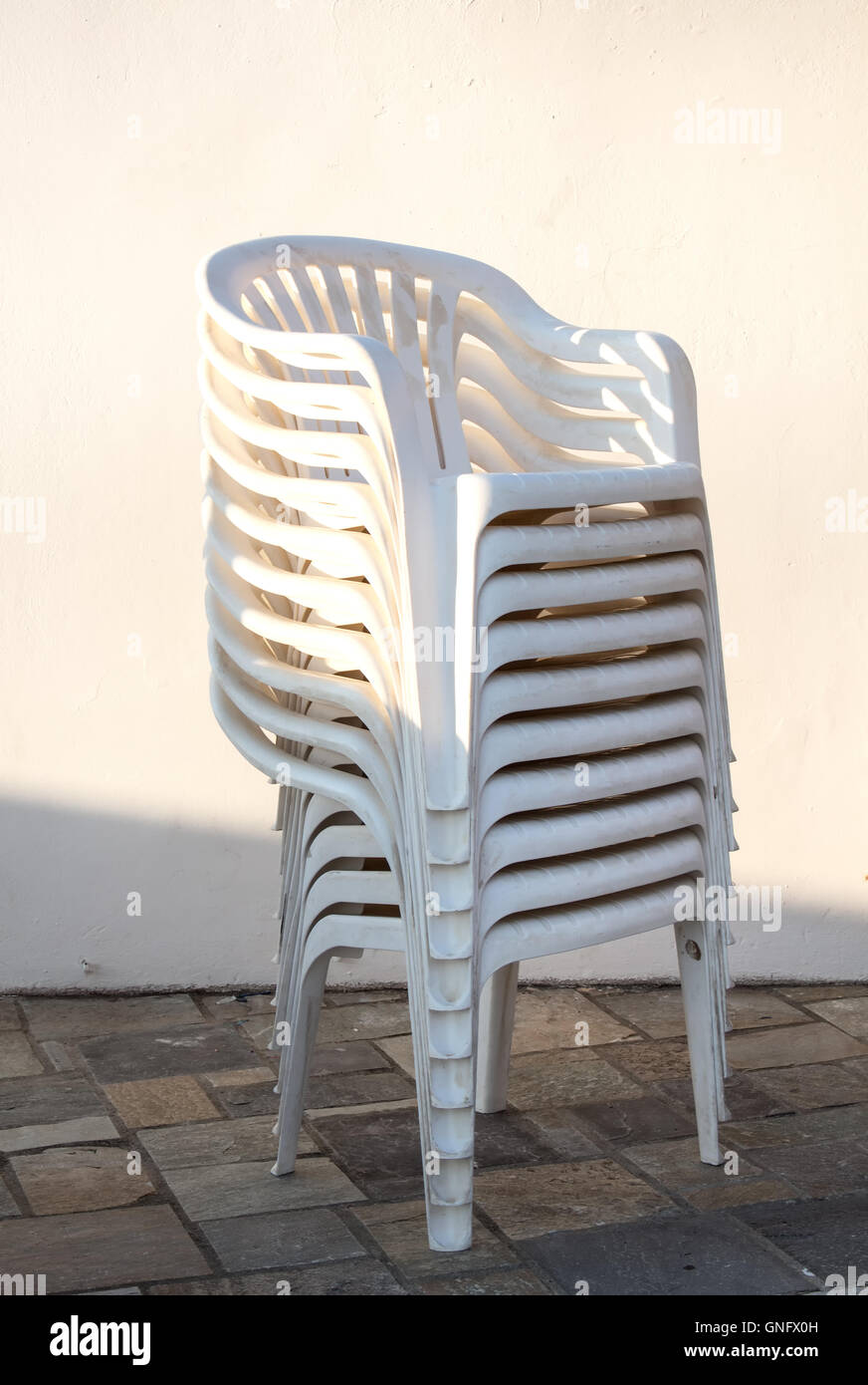Weiße Kunststoffstühle Avatara in einer Spalte. Stein Boden, weiße Wand im Hintergrund. Im Freien. Sonnenlicht am frühen Abend. Stockfoto