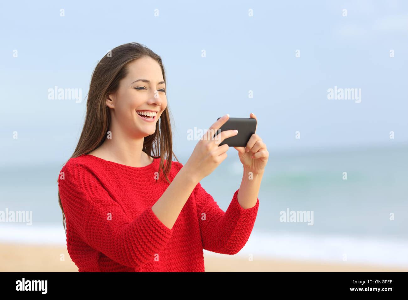 Porträt von ein fröhliches Mädchen mit roten Pullover lachen beim Ansehen von Streaming-Videos in Smartphone am Strand mit dem Meer Stockfoto