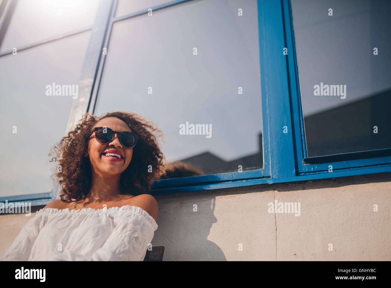 Schuss der glückliche junge afrikanische Frau sitzen im freien wegsehen und lächelnd. Frau mit Sonnenbrille draußen aussehende h Stockfoto